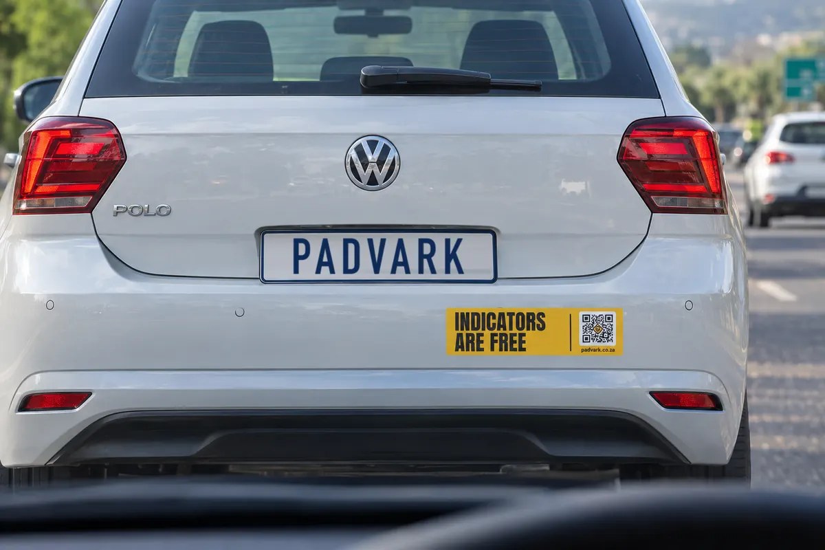 A white hatchback in traffic with the Indicators are free bumper sticker on its rear bumper