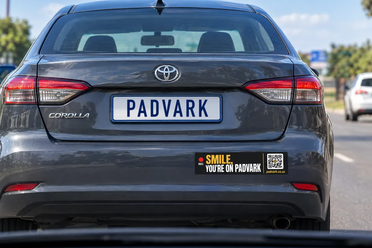 A grey sedan in traffic with the Smile, you're on Padvark bumper sticker on its rear bumper